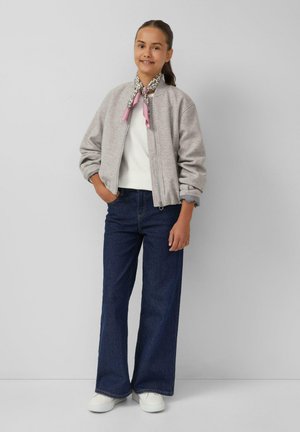 Jeune personne se tenant debout les mains dans les poches de sa veste, portant un sweat à zip gris, un haut blanc, un jean bleu foncé, des chaussures blanches et une écharpe à motifs.