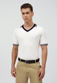 Polo de manga corta y cuello en V en blanco con rayas verticales, cuello y puños en azul marino. Combinado con pantalones caqui y cinturón negro.
