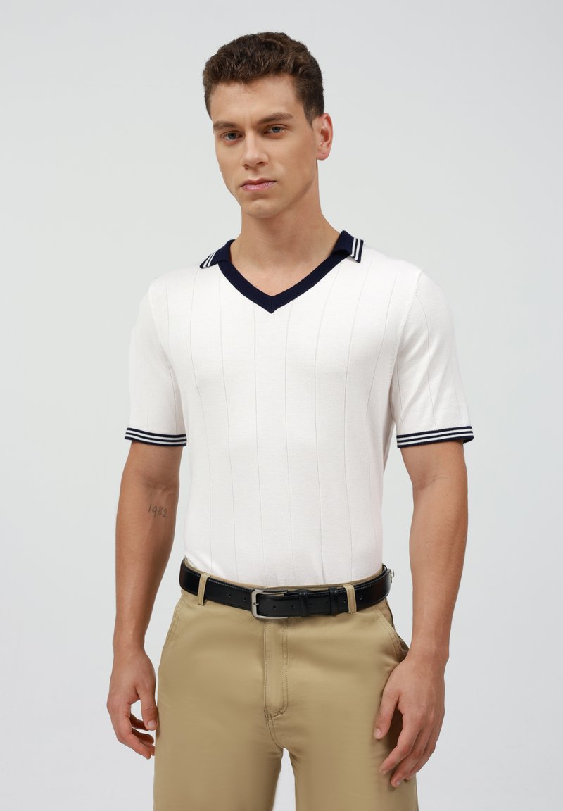 Polo de manga corta y cuello en V en blanco con rayas verticales, cuello y puños en azul marino. Combinado con pantalones caqui y cinturón negro.
