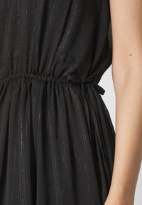 Robe noire en tissu texturé avec une lueur subtile. Taille froncée avec un détail de cordon. Silhouette fluide avec des plis.