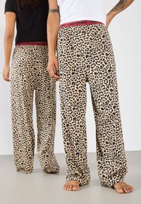 Pysjamasbukser med leopardmønster i beige med sorte prikker, med en rød elastisk talje med "URTRN" trykt. Løs, avslappet passform.
