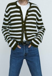 Henkilö, jolla on yllään valkoinen ja tummanvihreä raidallinen pörröinen kardigan valkoisen T-paidan päällä, yhdistettynä korkeavyötäröisiin sinisiin farkkuihin ja mustaan vyöhön.