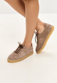 Beine mit beigen Wildleder-Schnürsneakers mit Sohlen aus Kaugummigummi, posierend vor einem schlichten hellen Hintergrund.