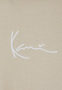 Țesătură bej cu o textură netedă, având un logo brodat alb cu scriptul "Kani" într-un stil fluid. Guler rotund, design minimalist.