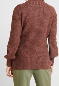 Žena v hnědém jemně strukturovaném pleteném svetru s dlouhým rukávem a zelených kalhotách, zobrazená zezadu na bílém pozadí.