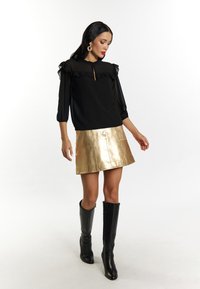Zwarte blouse met ruches, gouden metallic minirok en zwarte kniehoge laarzen. Glad textuur en gelaagd materiaal versterken de look.