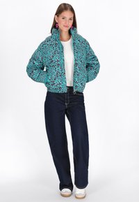 Jachetă puffer teal, cu imprimeu leopard în culori negre și roz, guler înalt, închidere cu fermoar, asortată cu un tricou deschis la culoare și blugi evazați de culoare albastru închis.