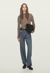 Frau mit welligem Haar trägt einen braunen Pullover, weite blaue Jeans, schwarzen Gürtel und dunkle Loafers, hält eine große, dunkelbraune Wildledertasche vor weißem Hintergrund.