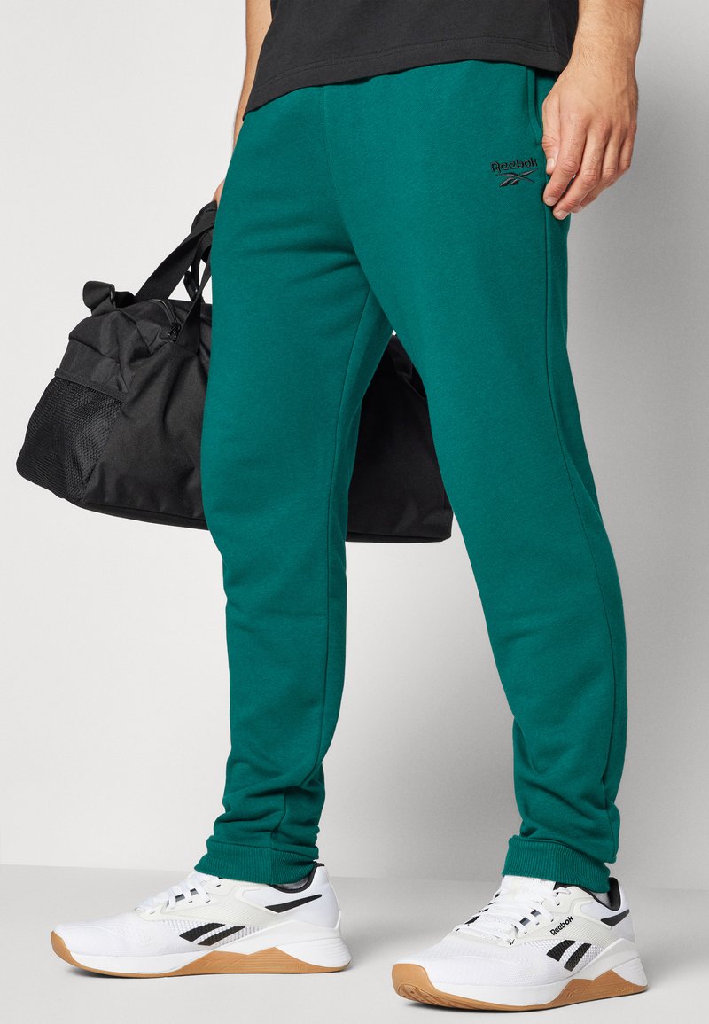 Pantalones deportivos verdes con cintura elástica, logo de Reebok en el costado, combinados con zapatillas deportivas en blanco y negro y una bolsa de deporte negra.