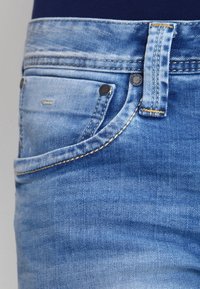 Ljust blå denimjeans med en klassisk femfickdesign, med guldtråd och svarta metallnitar på framsidan.