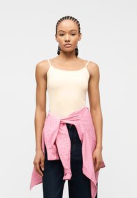 Νεαρή γυναίκα με κοτσίδες σινιόν, φοράει μπεζ μπλουζάκι χωρίς μανίκια, σκούρο τζιν και ροζ ριγέ πουκάμισο δεμένο στη μέση, μπροστά σε λευκό φόντο.