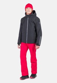 Giacca nera impermeabile con cappuccio, zip grigia, strisce riflettenti; pantaloni rossi brillanti; calzature nere con accenti rossi; tessuto testurizzato.