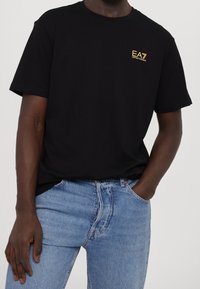 Camiseta de algodón negra con cuello redondo y mangas cortas, presenta el logo dorado de EA7 en el lado izquierdo del pecho, combinada con jeans de color azul claro.