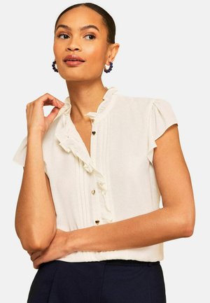 Frau in weißer Rüschenbluse mit herzförmigen Knöpfen und marineblauer Hose, trägt blaue Creolen, posiert mit Hand nahe am Gesicht.