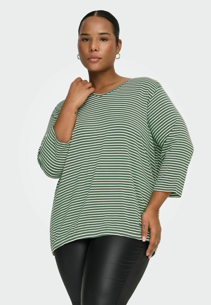 Kvinde med mørkt hår trukket tilbage iført grøn og hvidstribet langærmet trøje og sorte leggings, stående mod en ensfarvet baggrund.