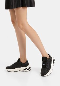 Sneakers atletici neri con suole bianche, caratterizzati da una parte superiore testurizzata, chiusura con lacci e marchi visibili sul tallone.