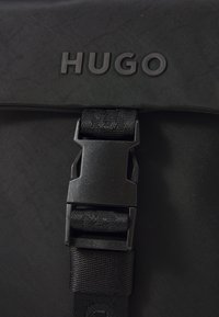 Svart ryggsekk med teksturert stoff, preget "HUGO"-logo, holdbar festelås og vevde stroppedetaljer.