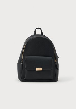 Mochila de couro preto com bolso frontal com aba e detalhes em zíper dourado, em pé sobre um fundo branco.