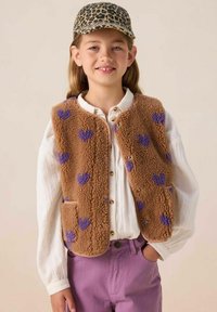 Gilet en peluche marron avec des motifs de cœurs violets, fermeture à boutons et deux poches, superposé sur un chemisier blanc, associé à un pantalon violet clair.