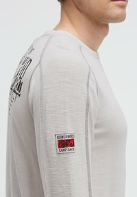 Hellgraues Langarmshirt aus strukturiertem Stoff, mit einem schwarzen Druck und einem rot-weißen Patch am linken Ärmel.