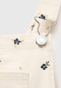 Beige Denim-Latzhose mit Blumenmuster und silbernem Druckknopfverschluss, verziert mit blauen und gelben Blumenmotiven an den Trägern und Taschen.