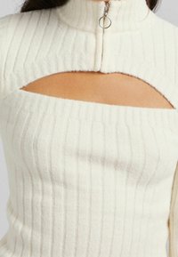 Vit ribbad stickad topp med hög krage och dragkedja samt en utskärning under kragen som skapar en figurnära silhuett.