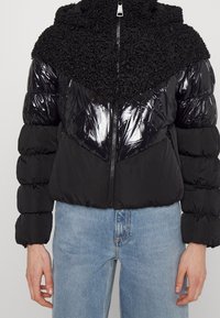 Svart pufferjacka med blank yta och texturerad päsliknande övre del. Dragkedjestängning, huva och samlade ärmar. Avslappnad design.