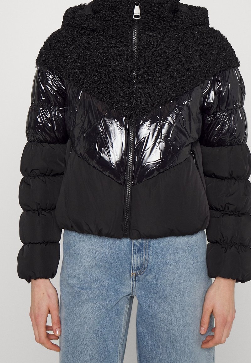 Svart pufferjacka med blank yta och texturerad päsliknande övre del. Dragkedjestängning, huva och samlade ärmar. Avslappnad design.