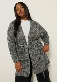 Kvinna med långt rakt hår som bär en svart- och vitrandig stickad cardigan över en vit topp och svarta byxor, poserar mot en enkel ljus vägg.