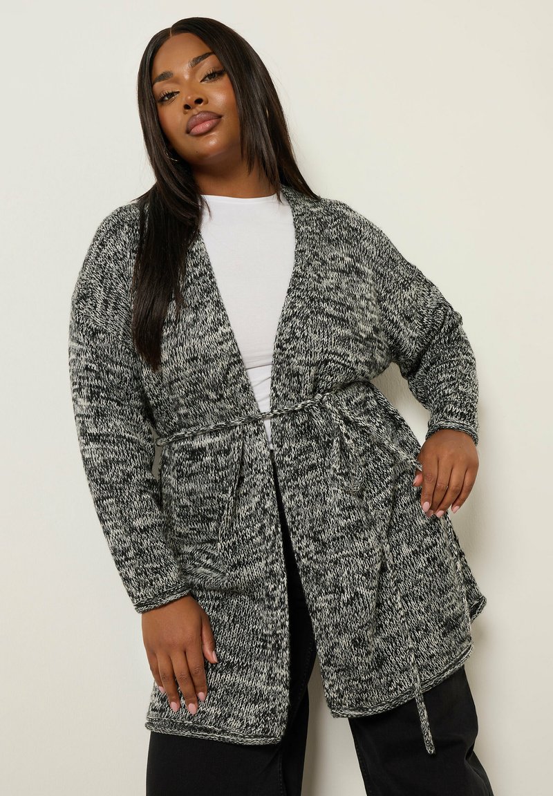 Kvinna med långt rakt hår som bär en svart- och vitrandig stickad cardigan över en vit topp och svarta byxor, poserar mot en enkel ljus vägg.