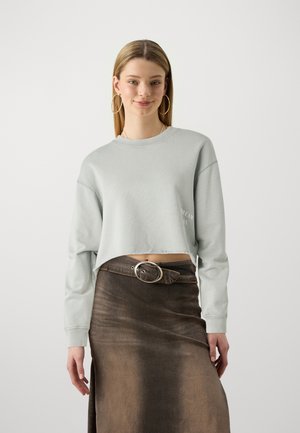 Lichtgrijze cropped sweatshirt met lange mouwen en ruwe zoom, gecombineerd met een bruine denimrok met een grote ronde gesp.