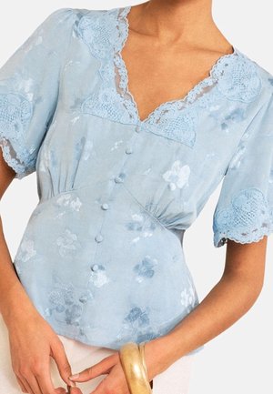 Blusa azul claro de manga corta con bordes de encaje floral y botones forrados de tela, llevada por una persona de piel clara que sostiene una pulsera rígida dorada.