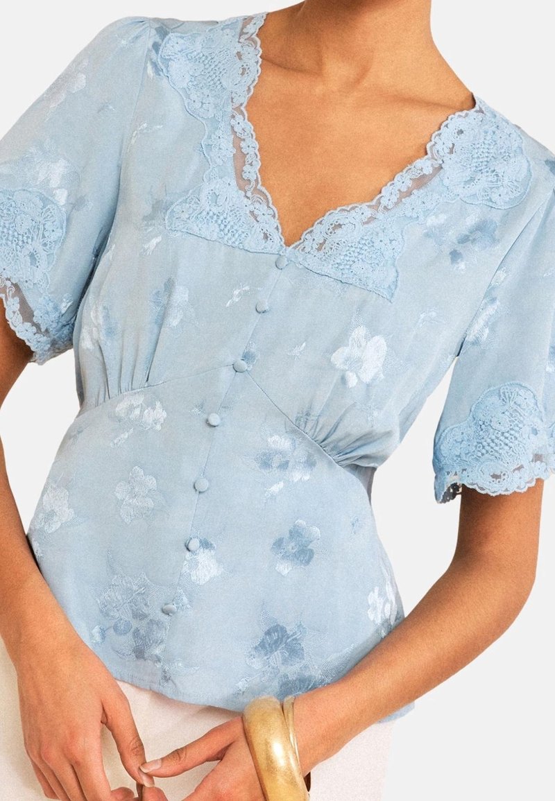 Blouse bleu clair à manches courtes avec bordure en dentelle florale et boutons recouverts de tissu, portée par une personne à la peau claire tenant un bracelet rigide en or.