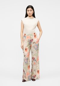 Kvinne med langt mørkt hår iført ermeløs kremfarget topp og vide bukser med blomsterprint i rosa, blått og grønt, stående med den ene hånden i lomma.