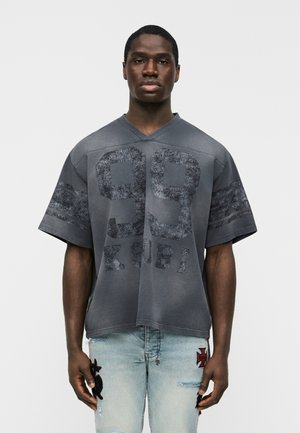 Homme portant une chemise ample gris foncé avec le numéro 99 et le texte "K SUBI", associée à un jean déchiré bleu clair avec des motifs en patch.
