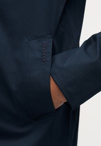 Σκούρο μπλε ναυτικό μανίκι και τσέπη ενός σακακιού με ένα χέρι μέσα στην τσέπη, με κεντημένο λογότυπο "Barbour" στην άκρη της τσέπης.