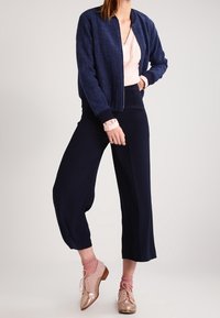 Marinefarbener, strukturierter Bomberjacke, rosa V-Ausschnitt-Top, weit geschnittene marinefarbene Hose und roségoldene Schnürschuhe, ergänzt durch rosa Socken.