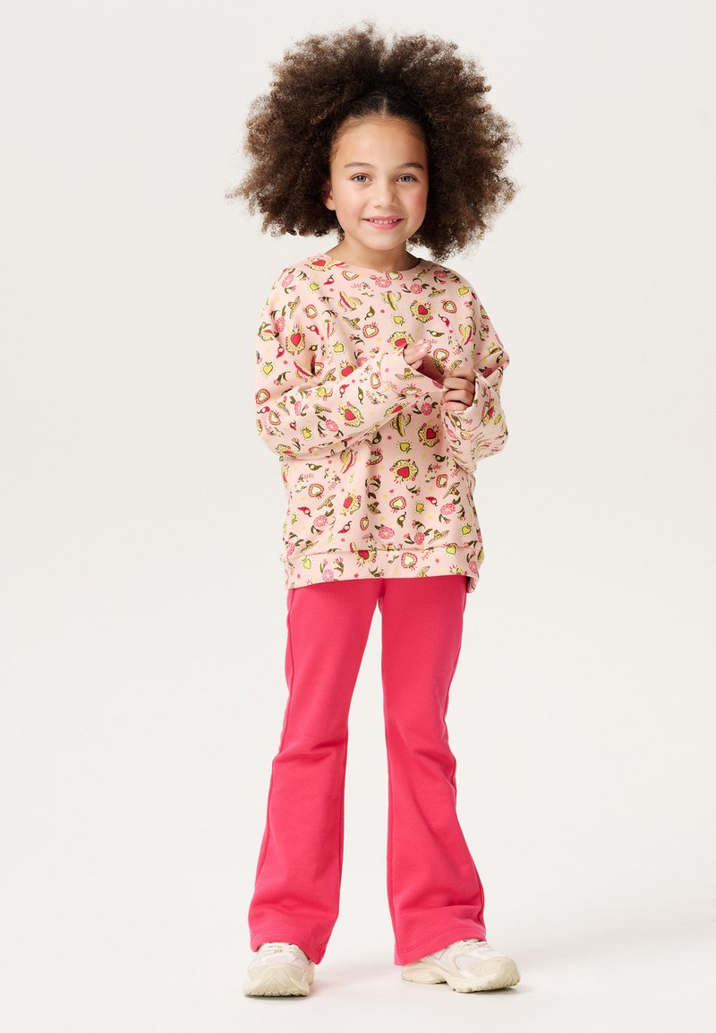 Sweat-shirt à manches longues rose clair avec des motifs de fruits et de fleurs colorés, associé à un pantalon évasé rose vif et des baskets blanches.