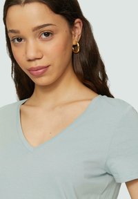 Helles blaues V-Ausschnitt-T-Shirt aus weichem Stoff mit kurzen Ärmeln, das eine glatte Textur und ein schlichtes Design aufweist. Es werden goldene Creolen getragen.