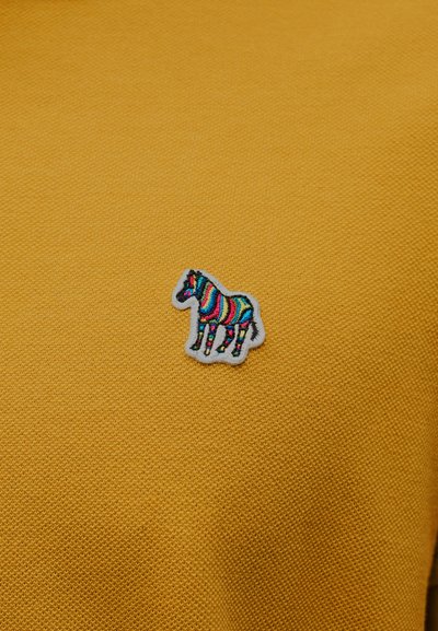 Κίτρινο υφασμάτινο με υφή που διαθέτει πολύχρωμο κεντημένο μπάλωμα ζέβρας με λευκό περίγραμμα, τοποθετημένο στην αριστερή πλευρά του ρούχου.