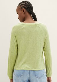 Grünes Langarmoberteil mit weichem Material, Raglanärmeln und rundem Ausschnitt, kombiniert mit hellblauen Jeans mit Gesäßtaschen.