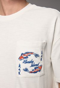 White cotton t-shirt featuring a left chest pocket with blue and orange embroidered koi fish and the text "Honshu Island."