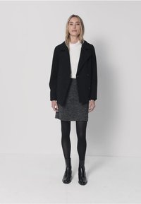 Manteau noir à double boutonnage porté sur un col roulé blanc, associé à une jupe grise texturée. Des bas noirs et des bottines complètent le look.