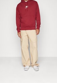 Bordeaux hoodie med en vit Nike-logga, beige cargobyxor och vita rutiga slip-on skor. Avslappnad passform med fickor på byxorna.