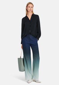 Marineblaues Button-Up-Hemd, fließende Ombre-Hosen, die von Dunkelblau zu Hellblau übergehen, und eine hellgrüne Tragetasche. Das Model trägt weiße spitze Schuhe.