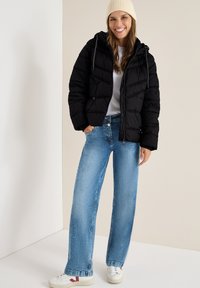Sort puffjakke med hætte, lynlås lukning og chevronmønster, parret med lyseblå vide jeans og hvide sneakers med røde accenter.