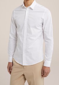 Man in een goed zittend wit overhemd met lange mouwen en beige broek, staand voor een effen achtergrond.