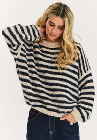 Sweter w paski z kremowymi i ciemnoniebieskimi pasami, o luźnym kroju, z okrągłym dekoltem i teksturowanym dzianym materiałem. Noszony z ciemnymi jeansami.