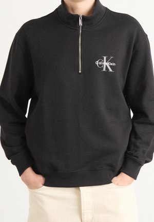 Personne portant un pull noir demi-fermeture éclair avec le logo Calvin Klein sur la poitrine et un pantalon crème, mains dans les poches.