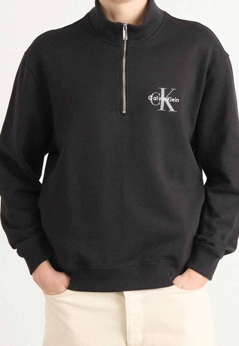 Personne portant un pull noir demi-fermeture éclair avec le logo Calvin Klein sur la poitrine et un pantalon crème, mains dans les poches.